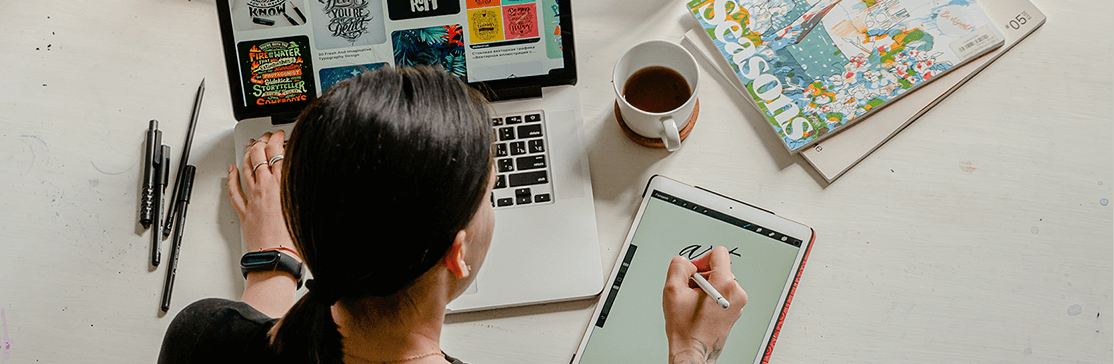 Woman sitting at white table at laptop, 4 pens to left of laptop, coffee and 2 magazines to the right, woman is writing on a tablet with her right hand and operating the laptop with her left