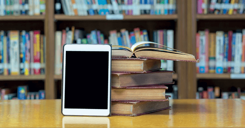 A tablet sitting on a library table leaning against 5 books. Full bookshelf in the background